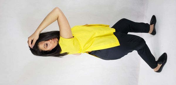 A woman with dark hair is wearing a sleeveless top cotton shirt with front back slits in bright yellow and black pants, seated on the floor against a plain light background, looking over her shoulder.