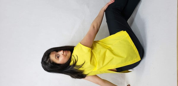 A woman with dark hair is wearing a sleeveless top cotton shirt with front back slits in bright yellow and black pants, seated on the floor against a plain light background, looking over her shoulder.