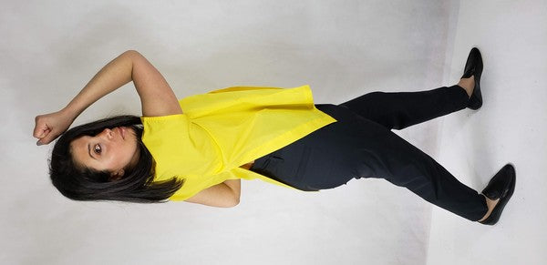 A woman with dark hair is wearing a sleeveless top cotton shirt with front back slits in bright yellow and black pants, seated on the floor against a plain light background, looking over her shoulder.