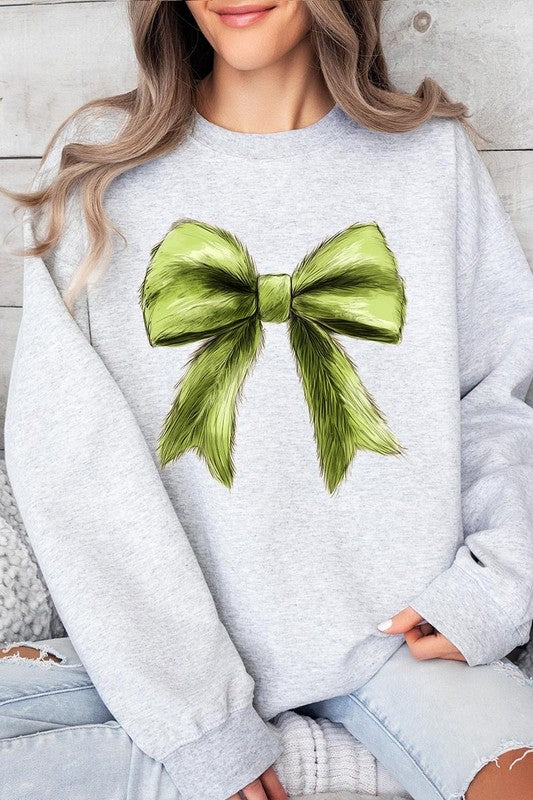 A person is wearing a green Grinchmas Coquette Bow Graphic Fleece Sweatshirt with a large, fluffy bow design on the front and blue distressed jeans. The background is a wooden wall.