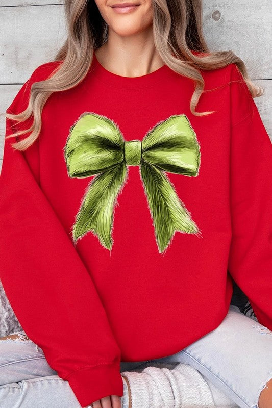 A person is wearing a green Grinchmas Coquette Bow Graphic Fleece Sweatshirt with a large, fluffy bow design on the front and blue distressed jeans. The background is a wooden wall.