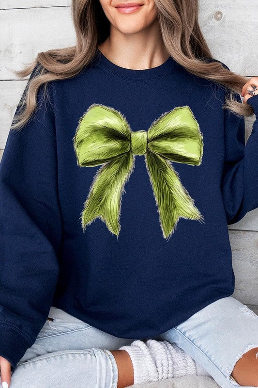 A person is wearing a green Grinchmas Coquette Bow Graphic Fleece Sweatshirt with a large, fluffy bow design on the front and blue distressed jeans. The background is a wooden wall.