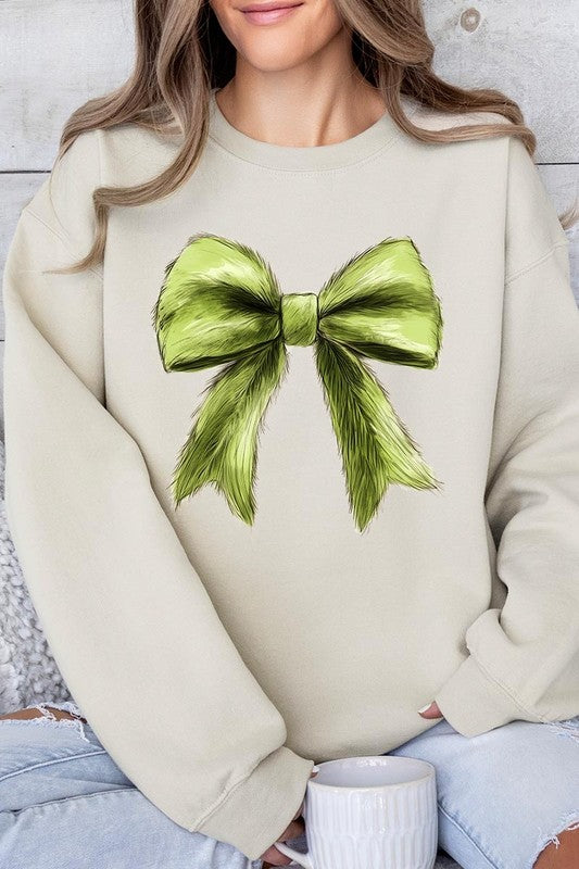 A person is wearing a green Grinchmas Coquette Bow Graphic Fleece Sweatshirt with a large, fluffy bow design on the front and blue distressed jeans. The background is a wooden wall.