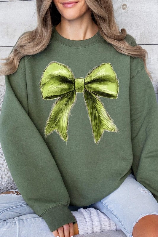 A person is wearing a green Grinchmas Coquette Bow Graphic Fleece Sweatshirt with a large, fluffy bow design on the front and blue distressed jeans. The background is a wooden wall.