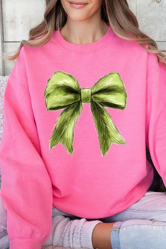 A person is wearing a green Grinchmas Coquette Bow Graphic Fleece Sweatshirt with a large, fluffy bow design on the front and blue distressed jeans. The background is a wooden wall.