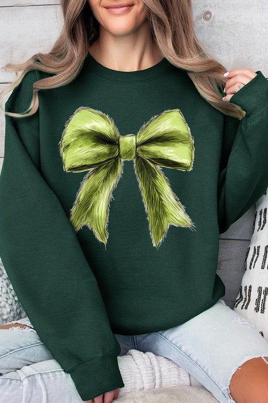 A person is wearing a green Grinchmas Coquette Bow Graphic Fleece Sweatshirt with a large, fluffy bow design on the front and blue distressed jeans. The background is a wooden wall.