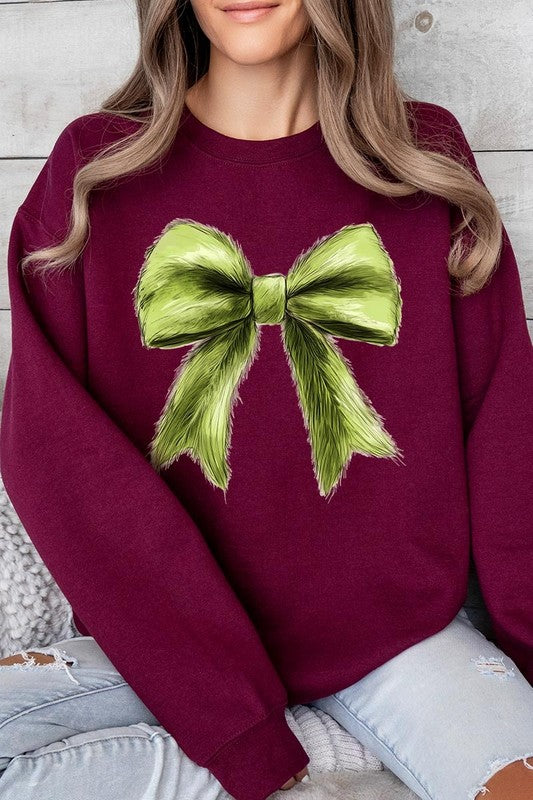 A person is wearing a green Grinchmas Coquette Bow Graphic Fleece Sweatshirt with a large, fluffy bow design on the front and blue distressed jeans. The background is a wooden wall.