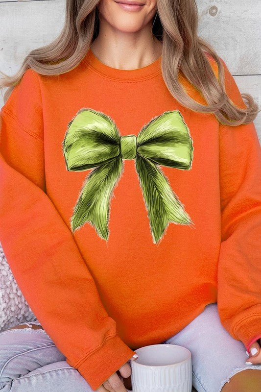 A person is wearing a green Grinchmas Coquette Bow Graphic Fleece Sweatshirt with a large, fluffy bow design on the front and blue distressed jeans. The background is a wooden wall.