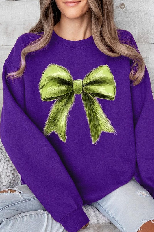 A person is wearing a green Grinchmas Coquette Bow Graphic Fleece Sweatshirt with a large, fluffy bow design on the front and blue distressed jeans. The background is a wooden wall.