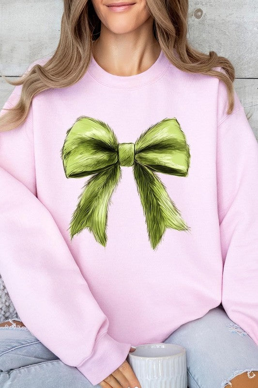 A person is wearing a green Grinchmas Coquette Bow Graphic Fleece Sweatshirt with a large, fluffy bow design on the front and blue distressed jeans. The background is a wooden wall.