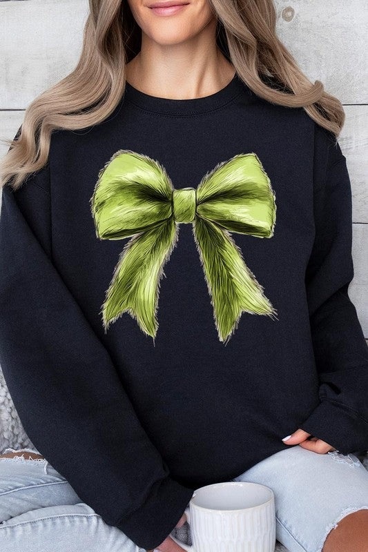 A person is wearing a green Grinchmas Coquette Bow Graphic Fleece Sweatshirt with a large, fluffy bow design on the front and blue distressed jeans. The background is a wooden wall.