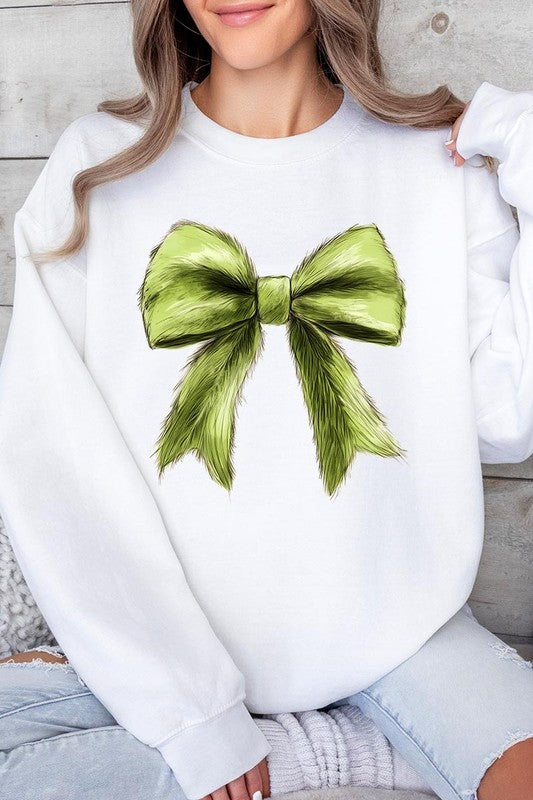 A person is wearing a green Grinchmas Coquette Bow Graphic Fleece Sweatshirt with a large, fluffy bow design on the front and blue distressed jeans. The background is a wooden wall.