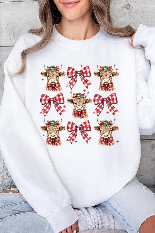 A person is wearing a Christmas Highland Cow Graphic Fleece Sweatshirt in light pink, showcasing a pattern of reindeer heads and red plaid bows.