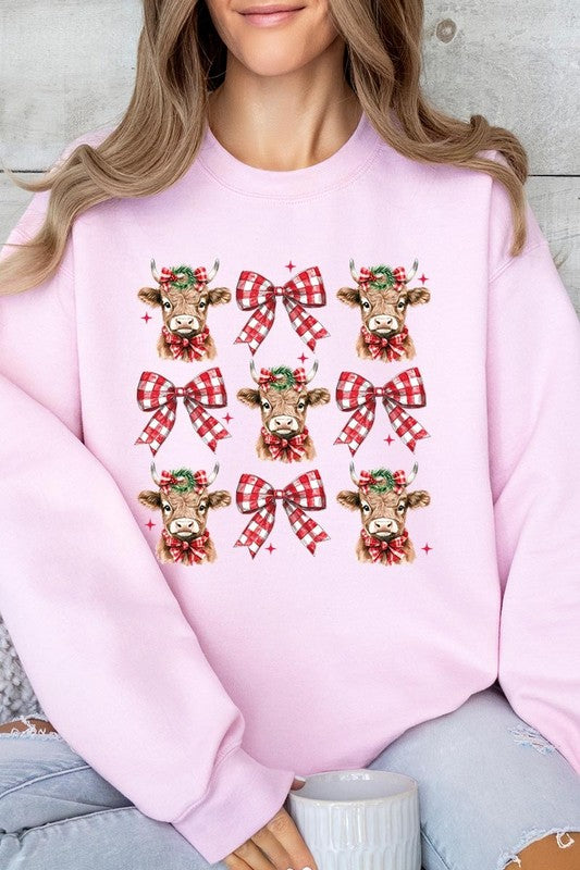 A person is wearing a Christmas Highland Cow Graphic Fleece Sweatshirt in light pink, showcasing a pattern of reindeer heads and red plaid bows.