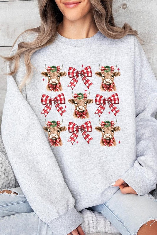 A person is wearing a Christmas Highland Cow Graphic Fleece Sweatshirt in light pink, showcasing a pattern of reindeer heads and red plaid bows.