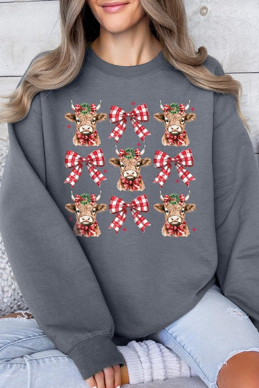 A person is wearing a Christmas Highland Cow Graphic Fleece Sweatshirt in light pink, showcasing a pattern of reindeer heads and red plaid bows.