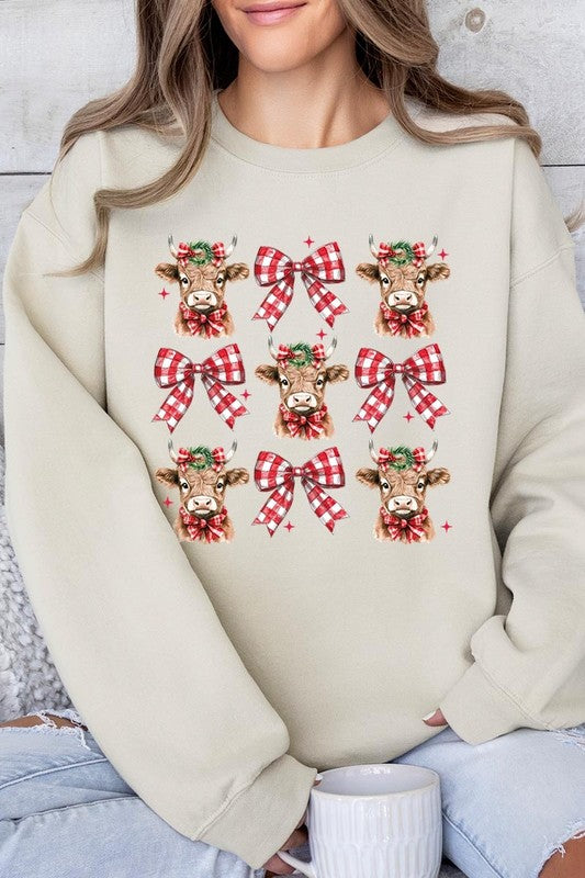 A person is wearing a Christmas Highland Cow Graphic Fleece Sweatshirt in light pink, showcasing a pattern of reindeer heads and red plaid bows.