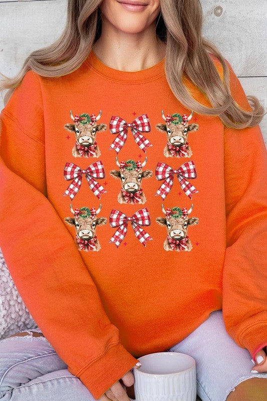 A person is wearing a Christmas Highland Cow Graphic Fleece Sweatshirt in light pink, showcasing a pattern of reindeer heads and red plaid bows.