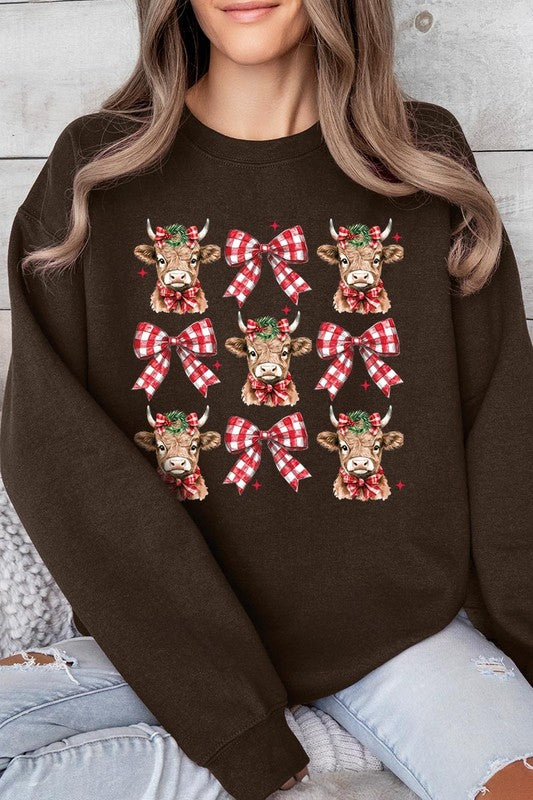 A person is wearing a Christmas Highland Cow Graphic Fleece Sweatshirt in light pink, showcasing a pattern of reindeer heads and red plaid bows.