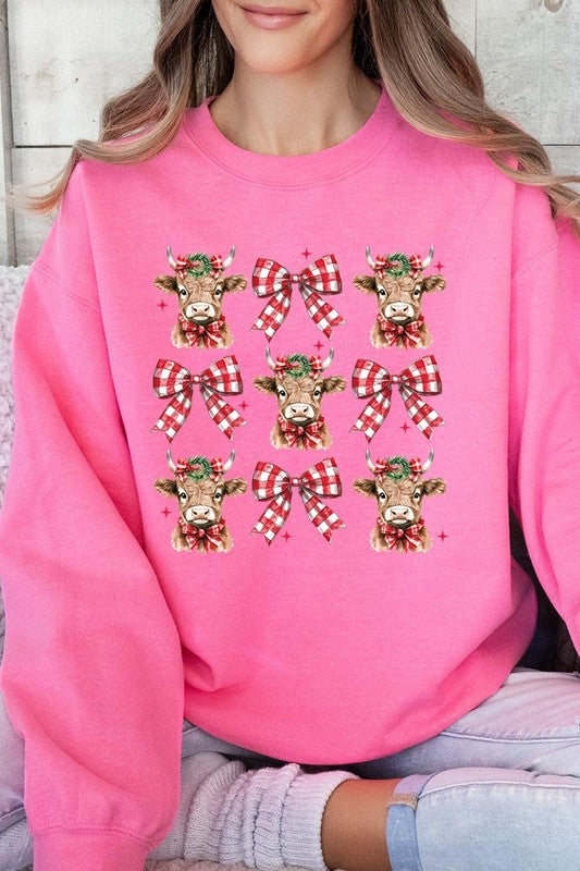 A person is wearing a Christmas Highland Cow Graphic Fleece Sweatshirt in light pink, showcasing a pattern of reindeer heads and red plaid bows.