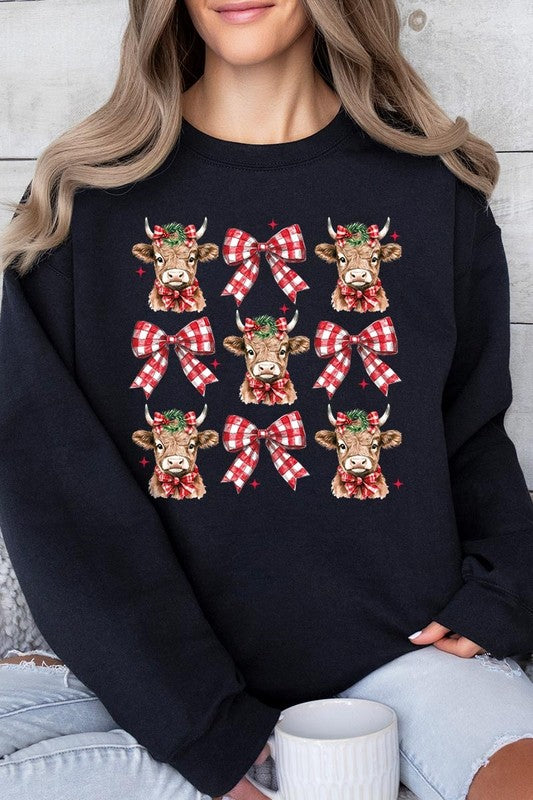 A person is wearing a Christmas Highland Cow Graphic Fleece Sweatshirt in light pink, showcasing a pattern of reindeer heads and red plaid bows.