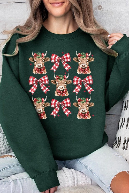 A person is wearing a Christmas Highland Cow Graphic Fleece Sweatshirt in light pink, showcasing a pattern of reindeer heads and red plaid bows.