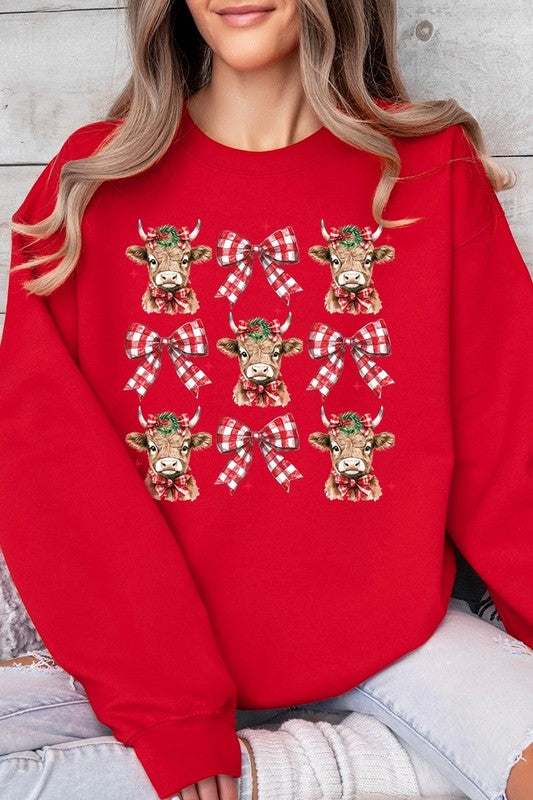 A person is wearing a Christmas Highland Cow Graphic Fleece Sweatshirt in light pink, showcasing a pattern of reindeer heads and red plaid bows.