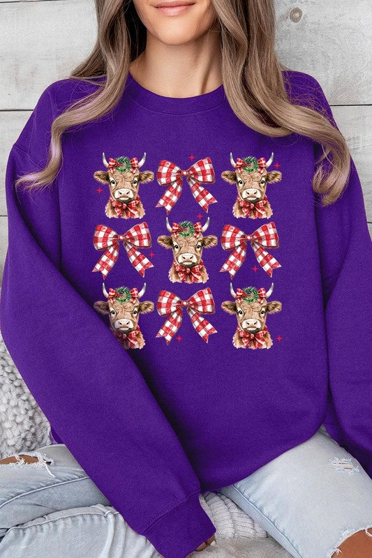 A person is wearing a Christmas Highland Cow Graphic Fleece Sweatshirt in light pink, showcasing a pattern of reindeer heads and red plaid bows.