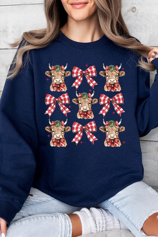 A person is wearing a Christmas Highland Cow Graphic Fleece Sweatshirt in light pink, showcasing a pattern of reindeer heads and red plaid bows.