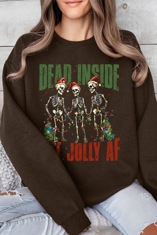 A person is wearing a "Dead Inside But Jolly AF Skeleton Sweatshirt," a beige unisex crew neck sweatshirt featuring a festive design of three dancing skeletons in Santa hats. The high-quality direct to film printed graphic text reads, "DEAD INSIDE BUT JOLLY AF.