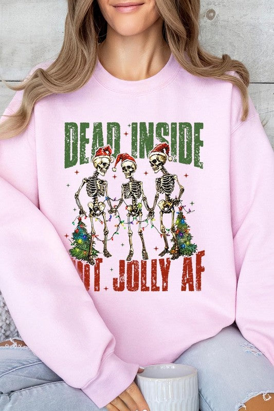 A person is wearing a "Dead Inside But Jolly AF Skeleton Sweatshirt," a beige unisex crew neck sweatshirt featuring a festive design of three dancing skeletons in Santa hats. The high-quality direct to film printed graphic text reads, "DEAD INSIDE BUT JOLLY AF.