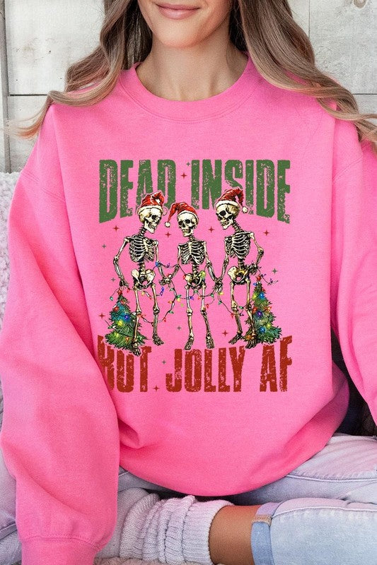 A person is wearing a "Dead Inside But Jolly AF Skeleton Sweatshirt," a beige unisex crew neck sweatshirt featuring a festive design of three dancing skeletons in Santa hats. The high-quality direct to film printed graphic text reads, "DEAD INSIDE BUT JOLLY AF.