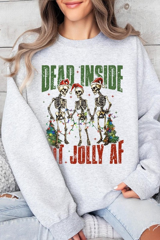 A person is wearing a "Dead Inside But Jolly AF Skeleton Sweatshirt," a beige unisex crew neck sweatshirt featuring a festive design of three dancing skeletons in Santa hats. The high-quality direct to film printed graphic text reads, "DEAD INSIDE BUT JOLLY AF.