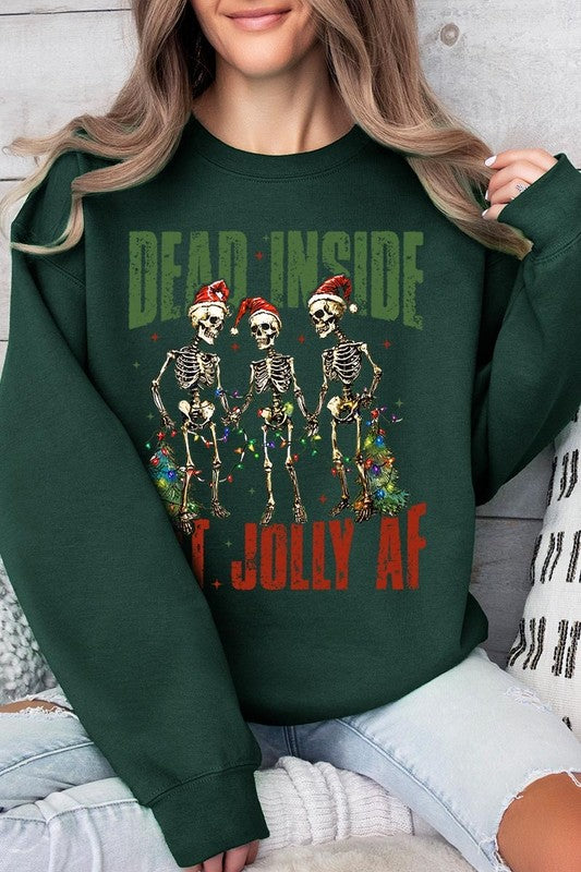 A person is wearing a "Dead Inside But Jolly AF Skeleton Sweatshirt," a beige unisex crew neck sweatshirt featuring a festive design of three dancing skeletons in Santa hats. The high-quality direct to film printed graphic text reads, "DEAD INSIDE BUT JOLLY AF.