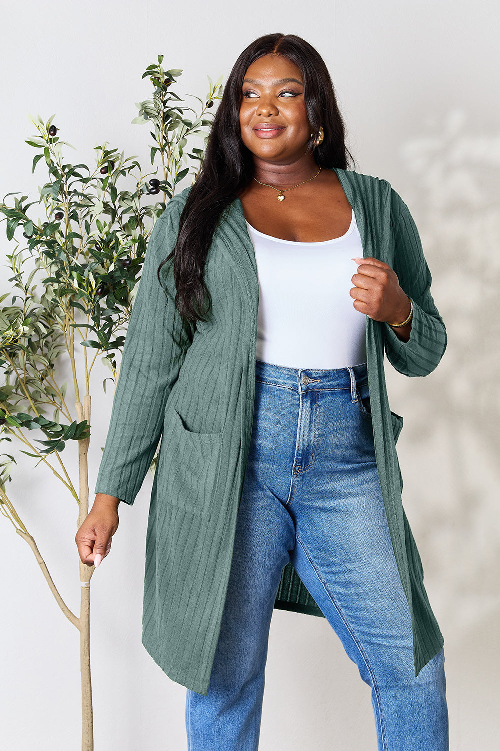 A woman wearing the Basic Bae Full Size Hooded Sweater Cardigan in black, paired with a white top and khaki pants, stands in front of a light background accented by a leafy plant on her left. With one hand tucked into her pocket, she gazes to her right.