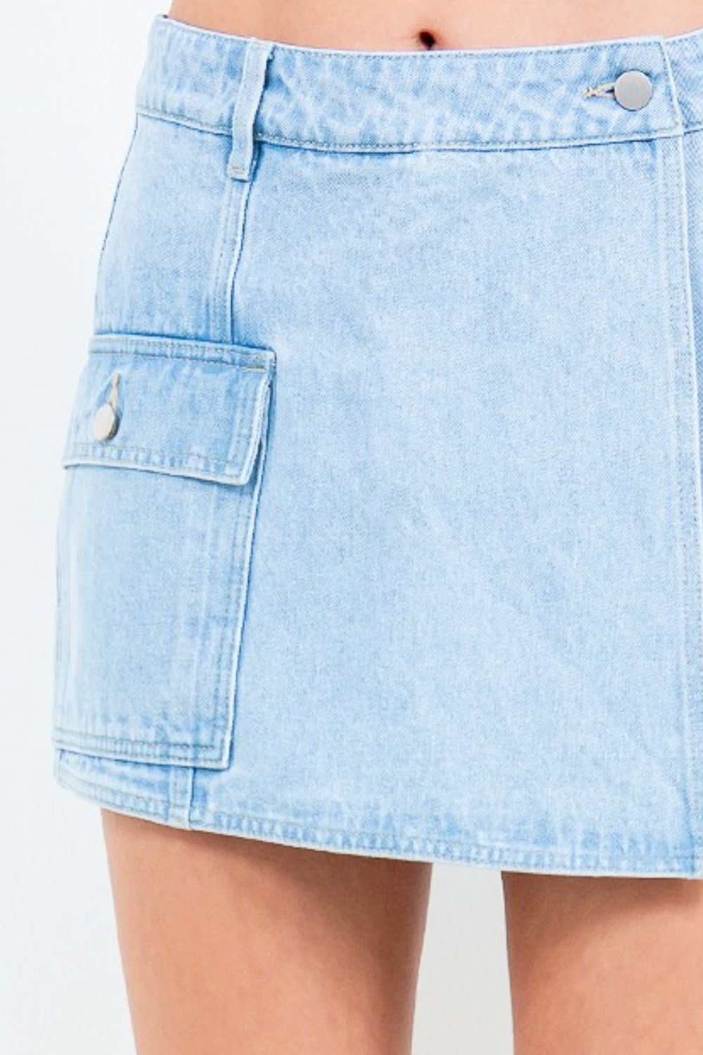 A person wears the American Bazi Cargo Pocket Mini Skort in light blue denim, featuring a large front pocket and button waist closure, paired with white sneakers.