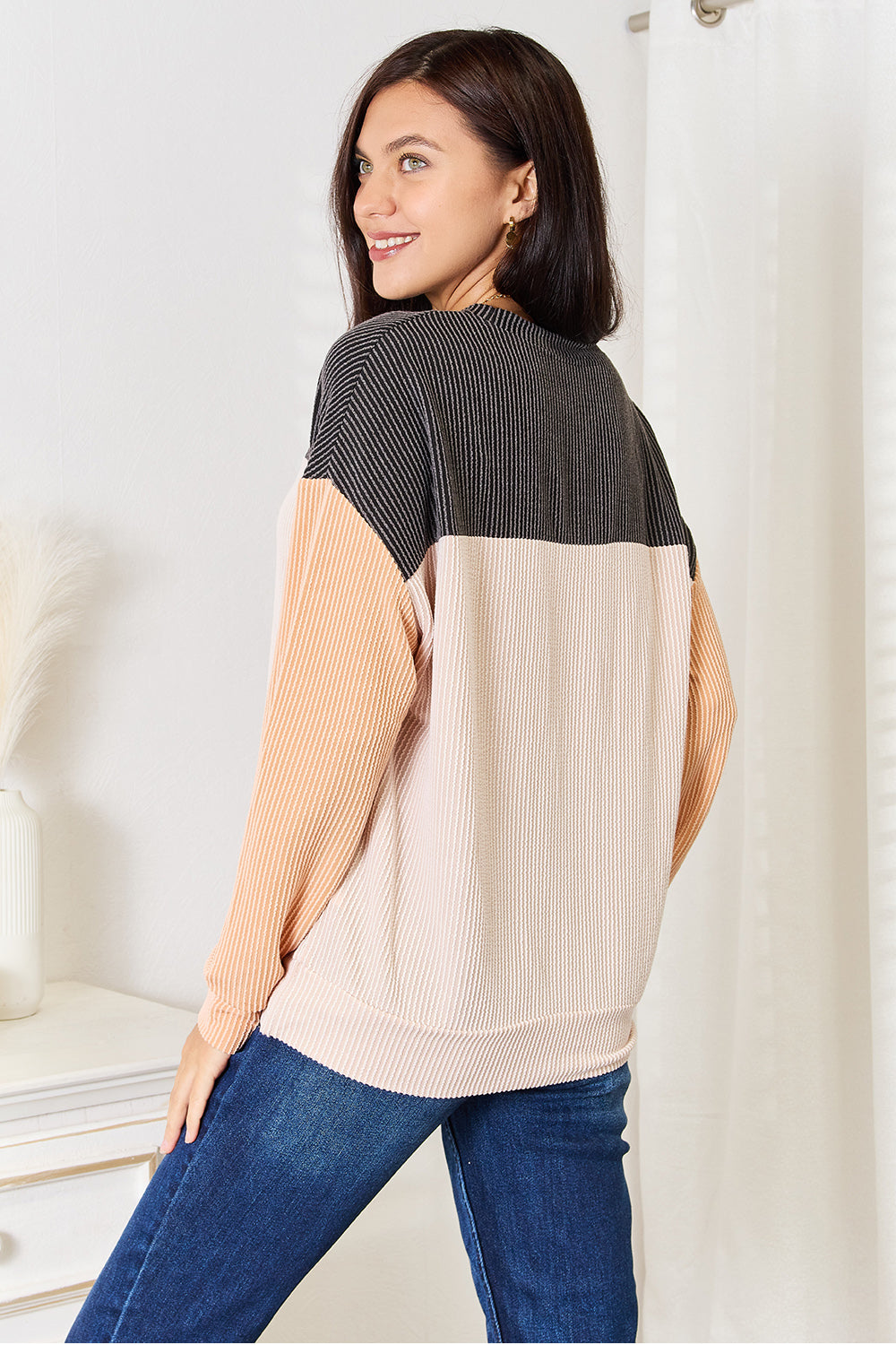 A woman with long brown hair stands indoors, smiling, and wearing a Double Take Color Block Dropped Shoulder T-Shirt paired with jeans. She is positioned near a small table with a white vase containing dried pampas grass, epitomizing the perfect casual wardrobe.