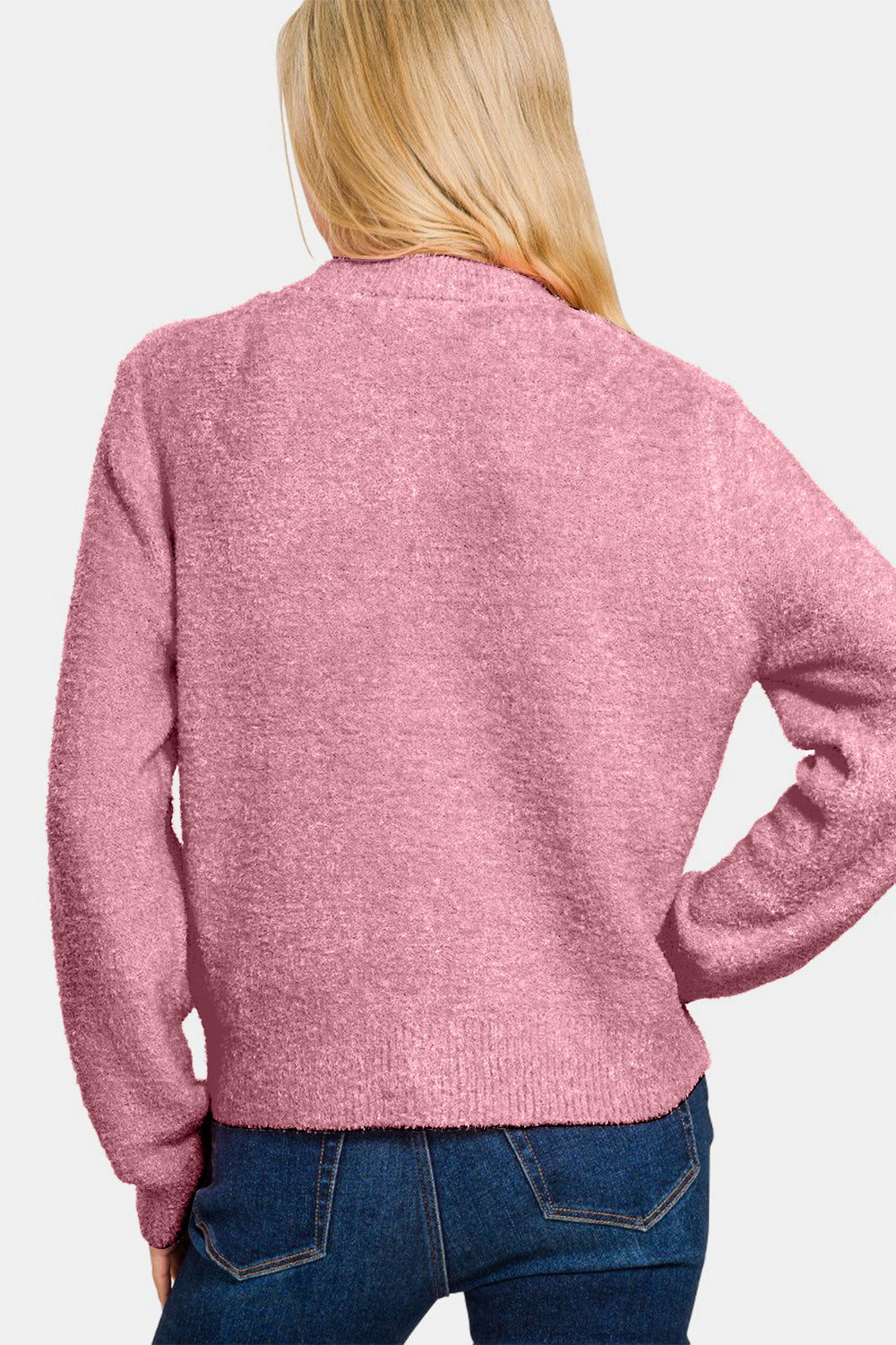 A person with long blonde hair is wearing a Zenana Button Down Long Sleeve Sweater Cardigan in pink over a white crop top and blue jeans, posing with one hand touching their hair.