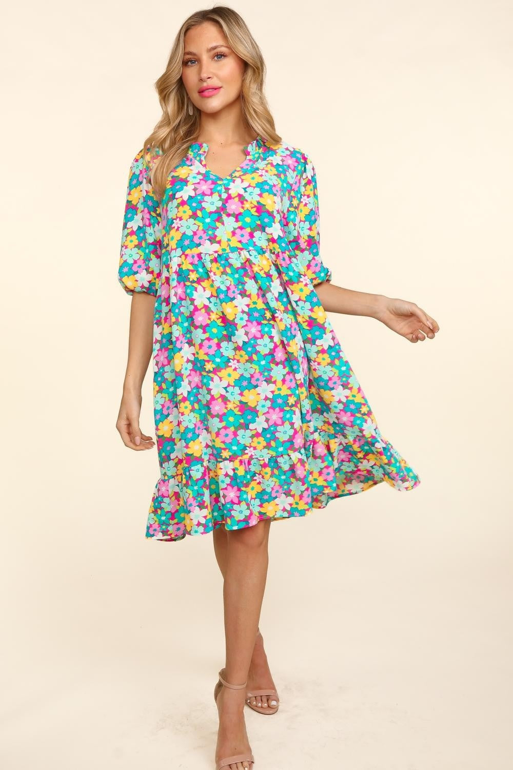 A woman in a Haptics Bubble Sleeve Floral Ruffled Dress stands against a plain background, with one hand holding her dress and the other by her side. She has long, wavy hair and is wearing open-toed shoes, perfect for any garden party.