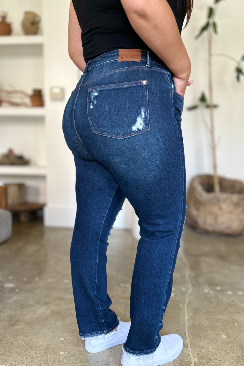 A woman in a black sleeveless top and Judy Blue Full Size High Waist Rigid Magic Heavy Destroy Straight Jeans stands indoors with her hands in her pockets. The background features a plant and shelves adorned with decor items.