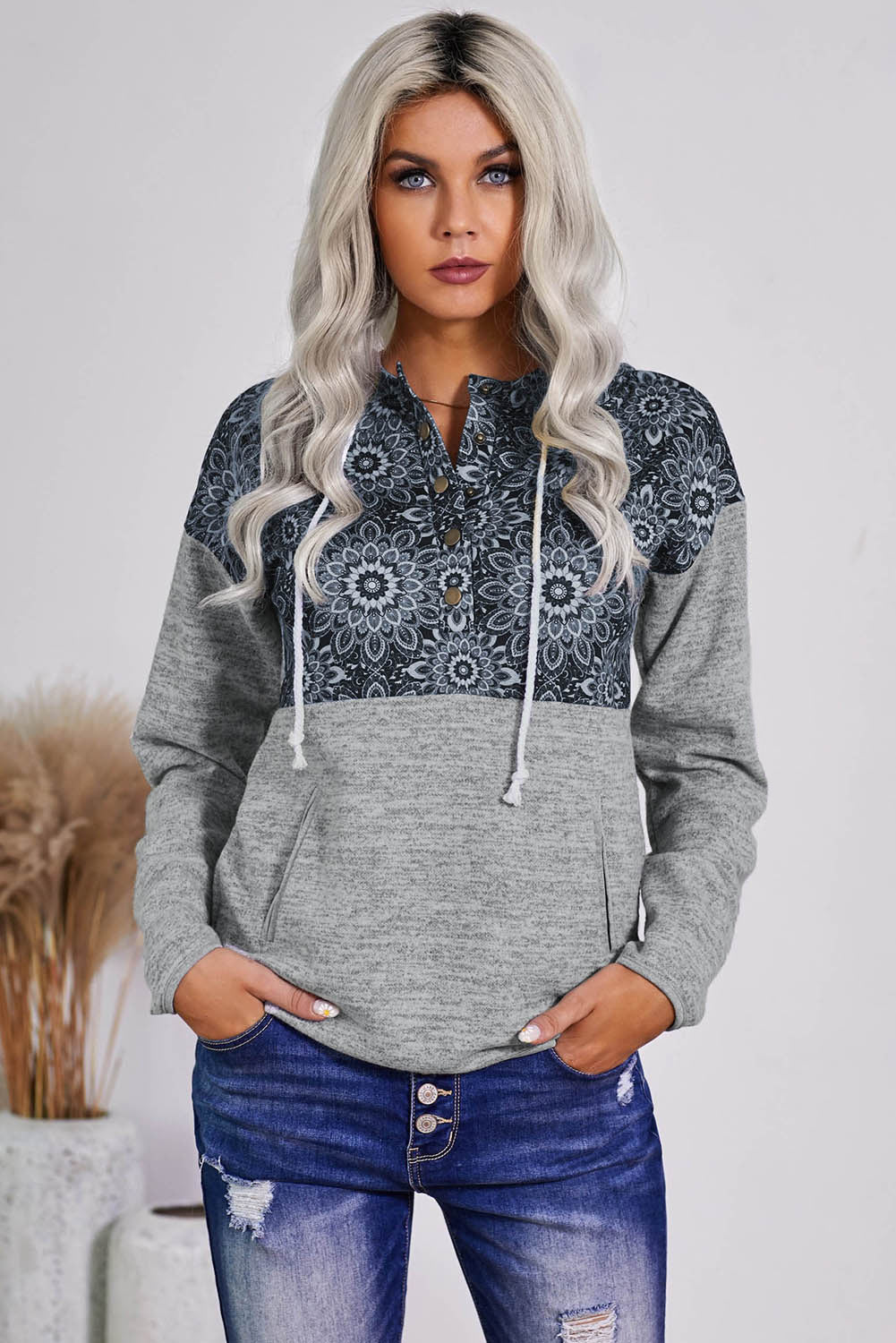 A person with long blonde hair is wearing a Gray Tribal Print Vintage Pocket Hoodie. They are turned sideways, and pampas grass in a vase is visible in the background.