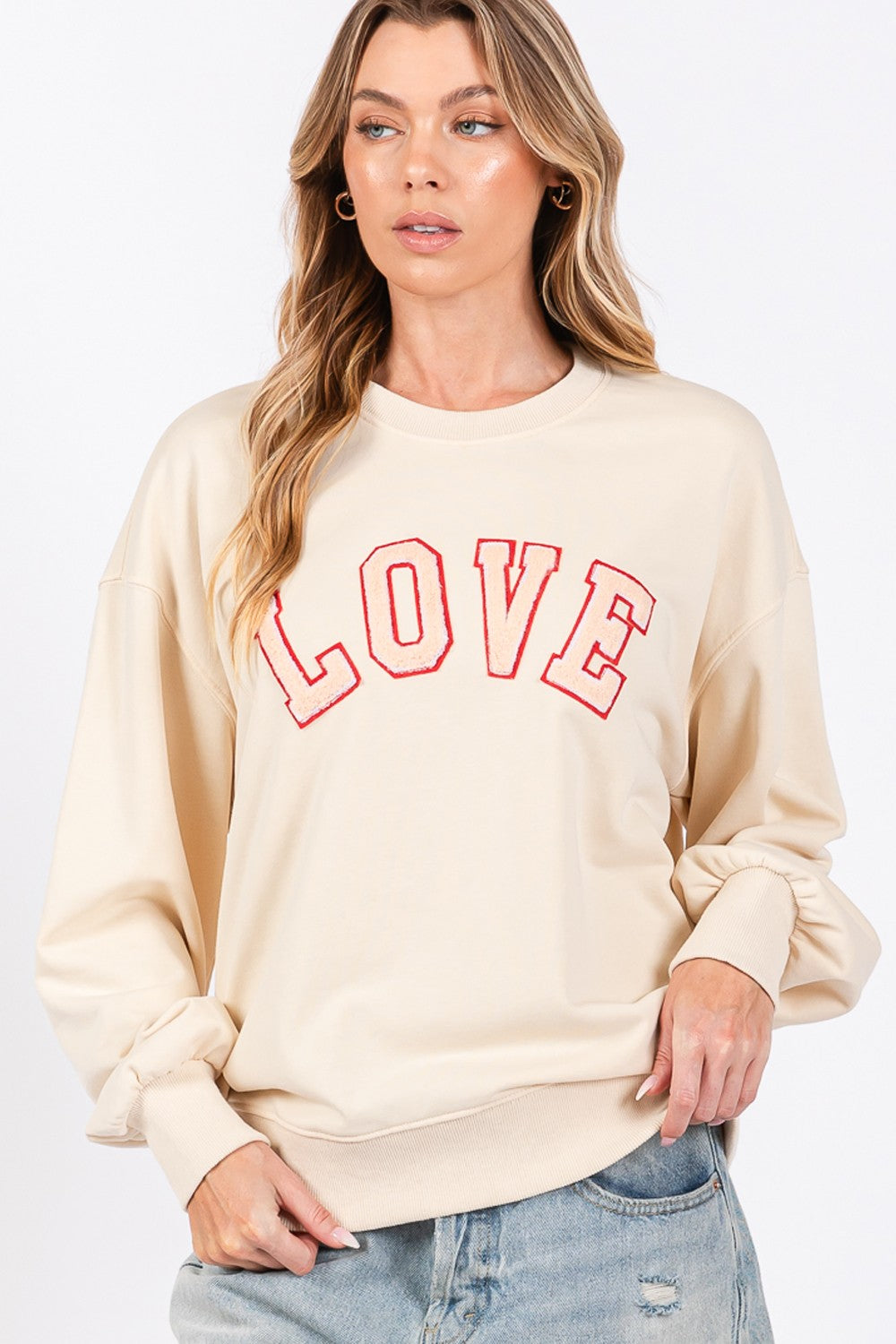 A woman dressed in a SAGE + FIG LOVE Path Applique Drop Shoulder Sweatshirt and a wide-brimmed hat is touching the brim of her hat with both hands, looking to the side and flaunting her cozy and stylish sweatshirt.