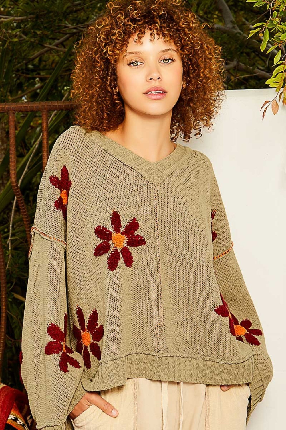 A person with curly hair wearing a fashionable outfit, featuring a POL V-Neck Floral Pattern Chenille Sweater in olive green adorned with red and orange flower patterns, is standing outdoors.