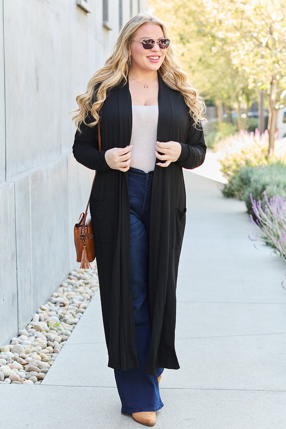 A woman stands against a concrete wall, wearing the Basic Bae Full Size Open Front Long Sleeve Cover Up made from rayon spandex, a white top, blue jeans in her perfect size, and brown shoes with her hands in her pockets. Machine wash cold for best results.