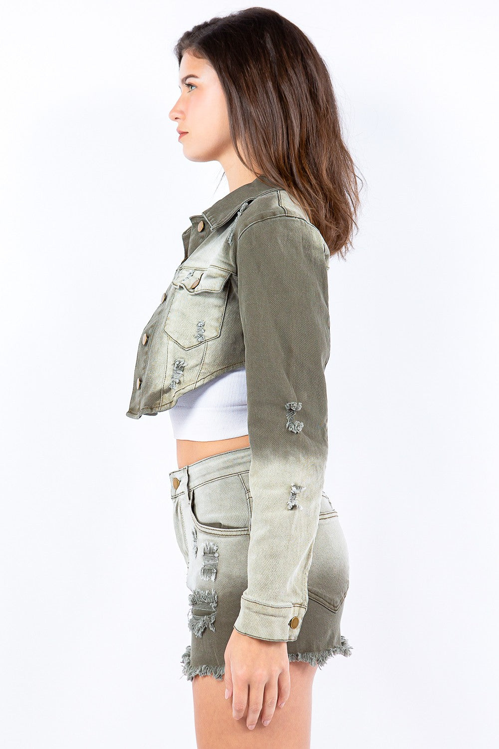 A person stands wearing an American Bazi Distressed Ombre Washed Cropped Denim Jacket in light olive green, paired with a matching skirt featuring a frayed hem and a white cropped tank top.