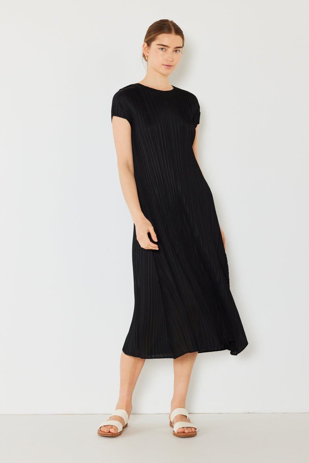 A person stands against a light wall, wearing an elegant Marina West Swim Pleated Cap Sleeve A-Line Dress in black and white sandals.