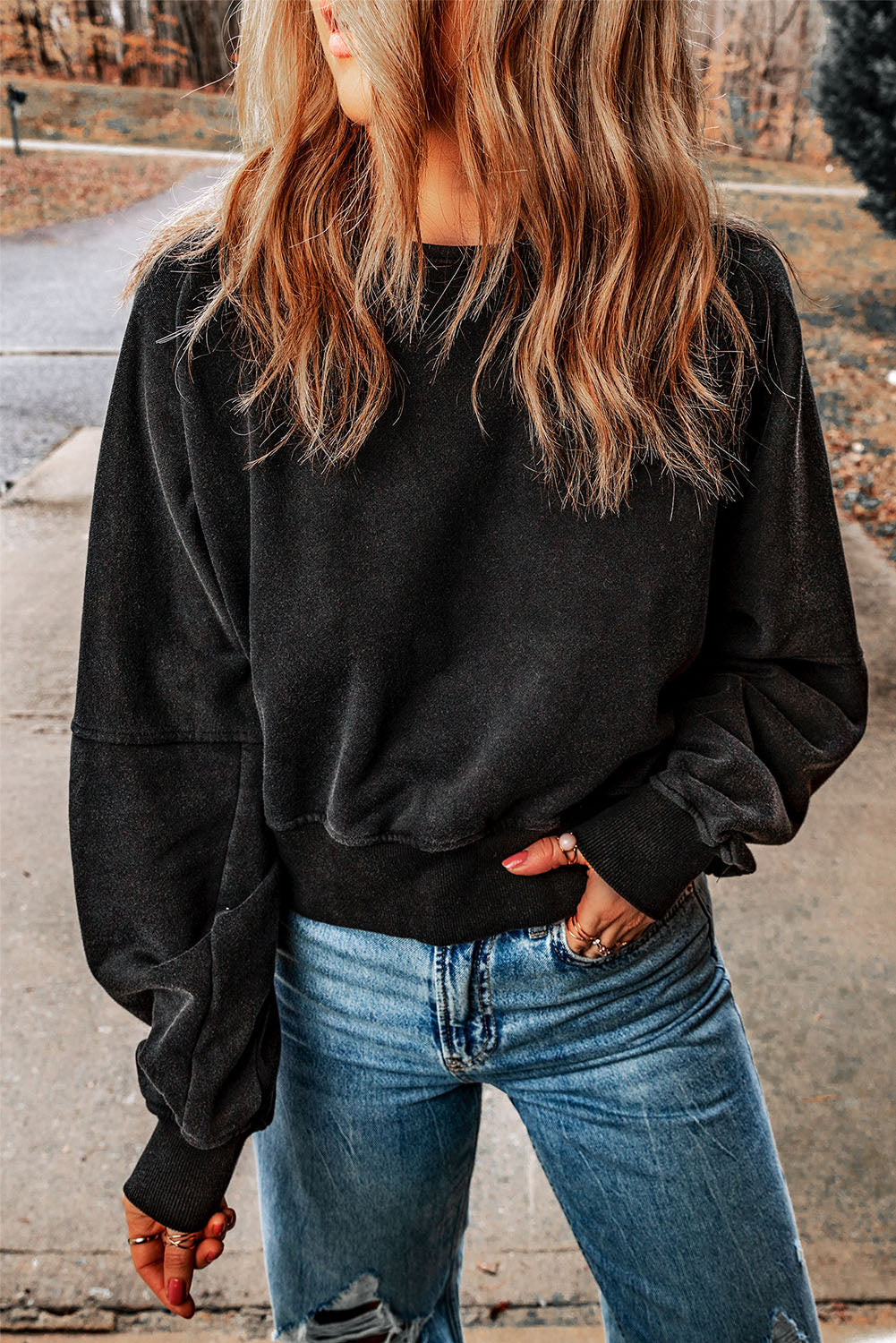 A person with wavy, shoulder-length hair wearing a Black Acid Wash V-shape Open Back Sweatshirt and distressed blue jeans stands outside on a paved surface.