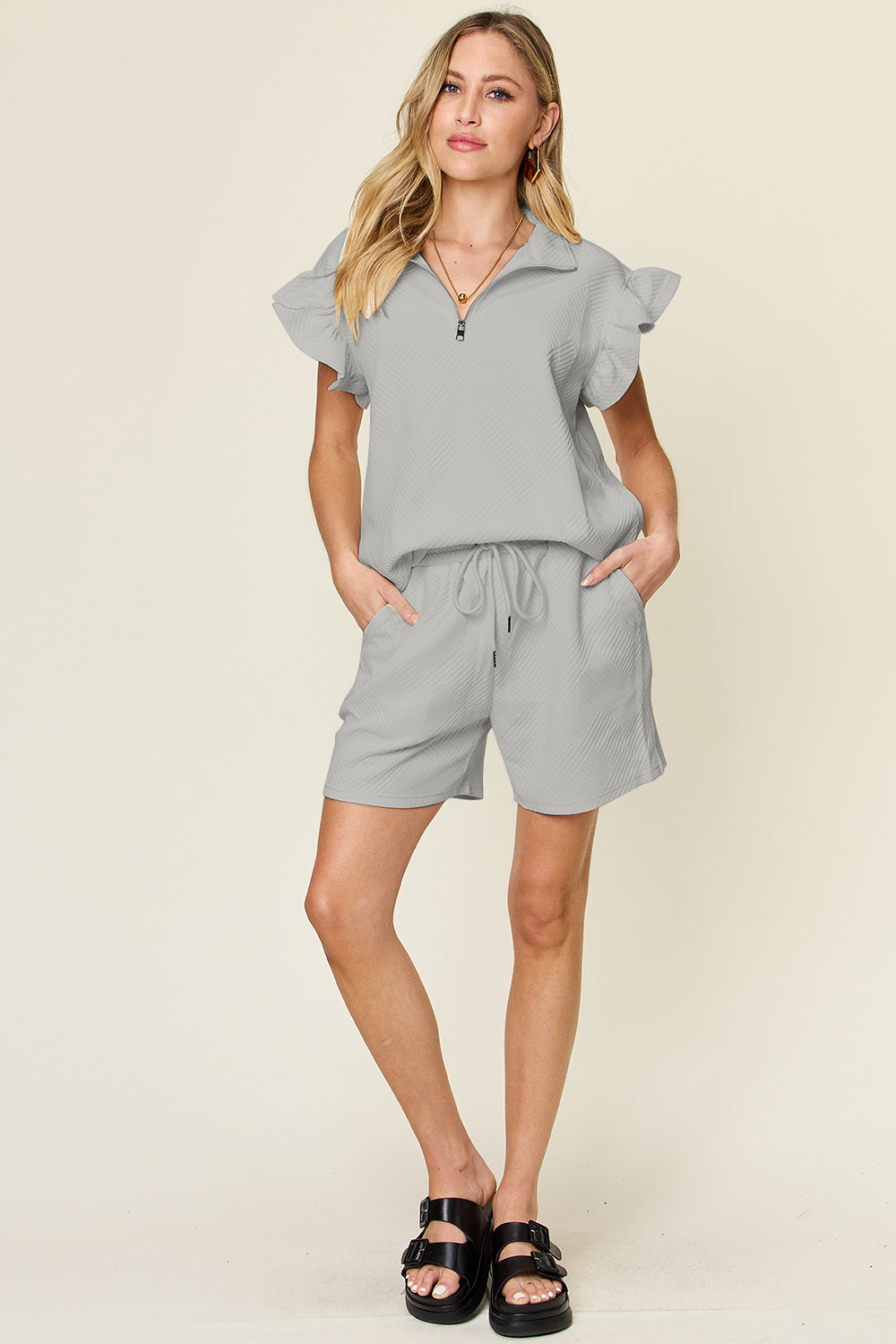 Against a plain background, a woman with blonde hair poses confidently with one hand in her pocket, highlighting the chic style of her Double Take Full Size Texture Flounce Sleeve Top and Drawstring Shorts Set.