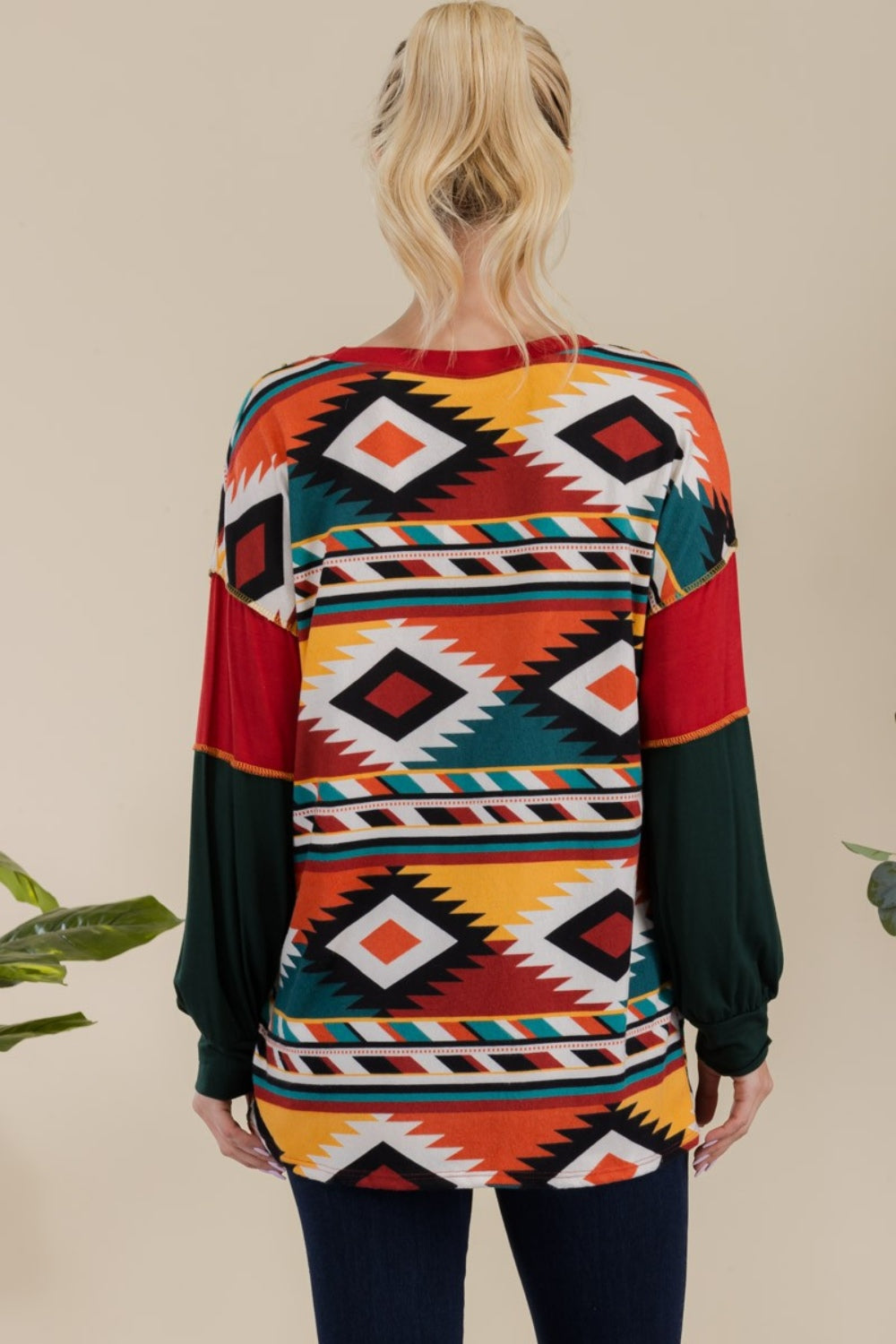 A woman stands against a plain background, wearing the Celeste Full Size Exposed Seam Printed Color Block T-Shirt with vibrant red, green, and yellow geometric sections, paired with dark jeans. She smiles, her hands resting in her jeans' pockets.