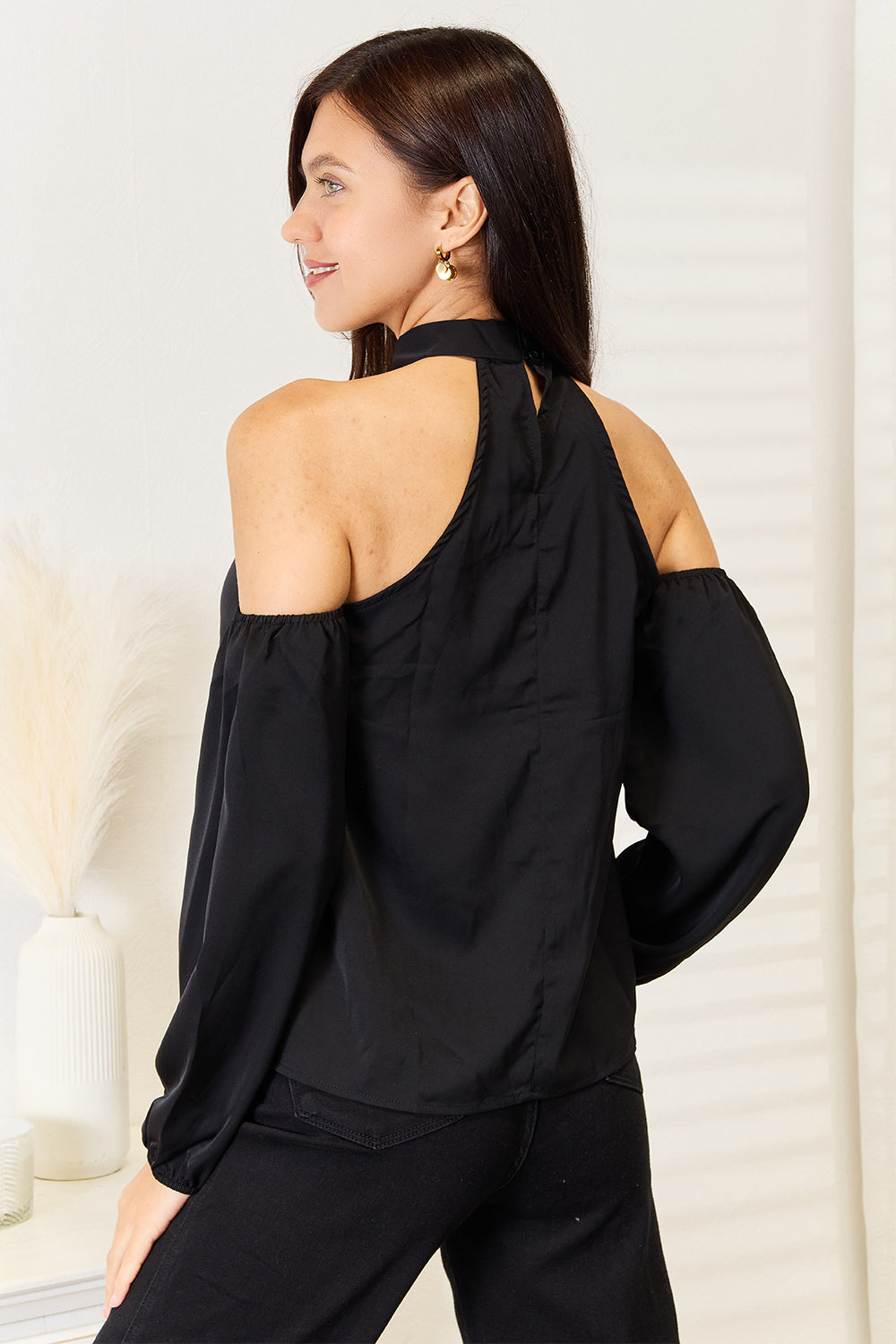 A woman with long brown hair wearing a Perfee Grecian Cold Shoulder Long Sleeve Blouse stands in a brightly lit room next to a white vase with pampas grass. She looks to the side with a smile.